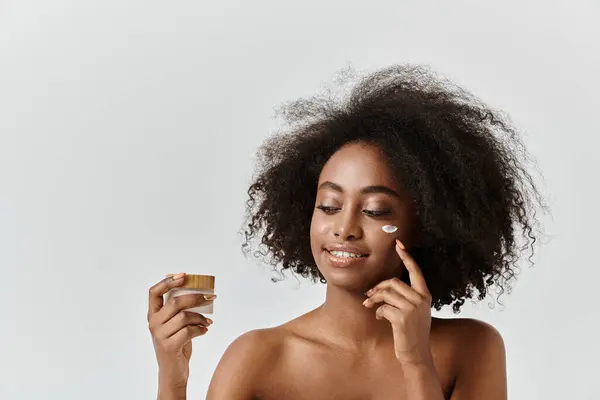 Una hermosa joven afroamericana con el pelo rizado sosteniendo un frasco de crema - foto de stock