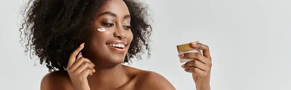 A beautiful young African American woman with curly hair applies cream on her face, focusing on skin care. — Stock Photo