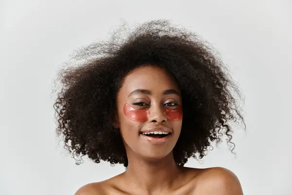 Une belle jeune femme afro-américaine aux cheveux bouclés présentant des patchs oculaires dans un studio, incarnant un concept de soins de la peau. — Photo de stock