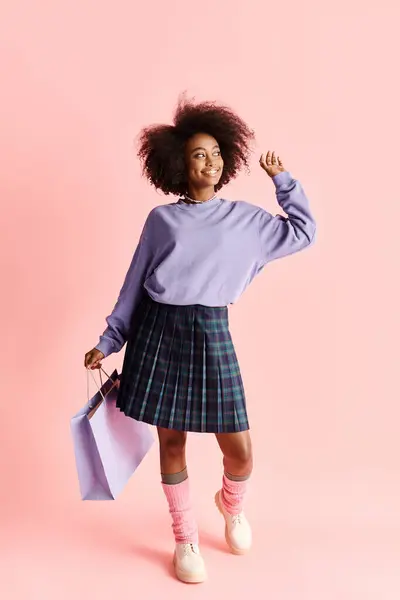 A fashionable African American woman with curly hair holds shopping bags in a purple sweater and plaid skirt. — Stock Photo
