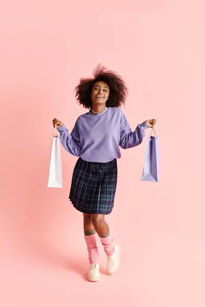 A young African American woman with curly hair, wearing a purple sweater and plaid skirt, holds shopping bags in a stylish studio photo shoot. — Stock Photo
