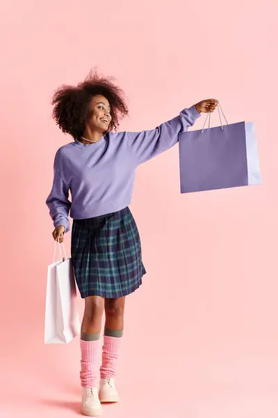 Una joven afroamericana con el pelo rizado sonriendo mientras sostiene bolsas llenas de compras con estilo. - foto de stock