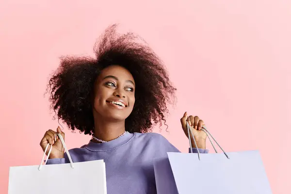 Una joven afroamericana con el pelo rizado sonriendo mientras sostiene bolsas de compras en un ambiente de estudio. - foto de stock