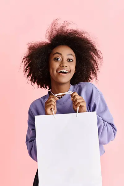 A beautiful young African American woman with curly hair holding a shopping bag and smiling in a studio setting. — Stock Photo