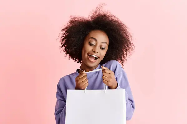 A beautiful young African American woman with curly hair smiling while holding a shopping bag in a studio setting. — Stock Photo