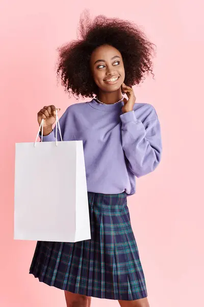 Uma jovem afro-americana com cabelo encaracolado está elegantemente em uma saia, segurando um saco de compras cheio de itens de moda. — Fotografia de Stock