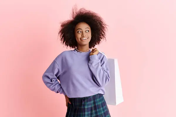 Una hermosa joven afroamericana con el pelo rizado en un suéter púrpura y falda a cuadros sosteniendo una bolsa de compras. - foto de stock