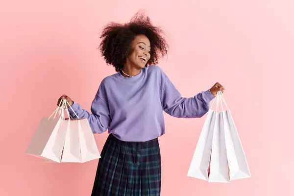 Young African American woman with curly hair, wearing a purple sweater and plaid skirt, holds shopping bags in studio. — Stock Photo