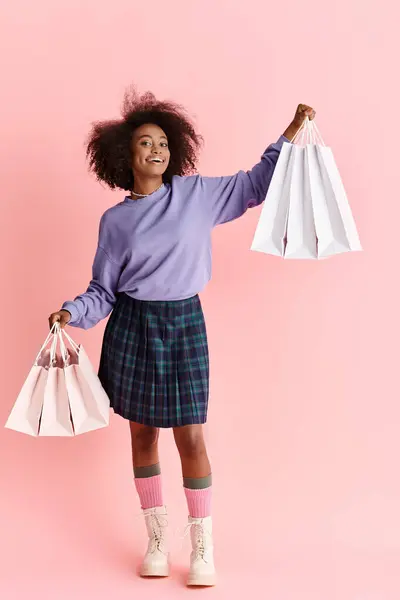 Giovane donna afroamericana con i capelli ricci indossa un maglione viola e gonna a quadri, tiene borse della spesa. — Foto stock