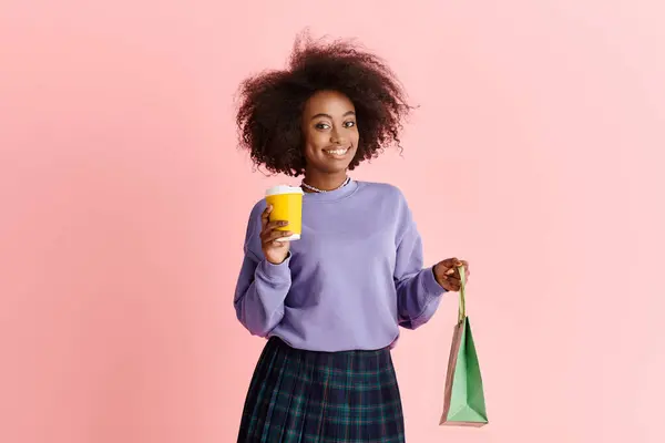 A stylish African American woman with curly hair holds a cup of coffee and a paper bag in a fashionable studio setting. — Stock Photo