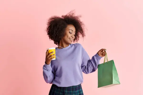 A young African American woman with curly hair holding a cup of coffee and a shopping bag in a studio setting. — Stock Photo