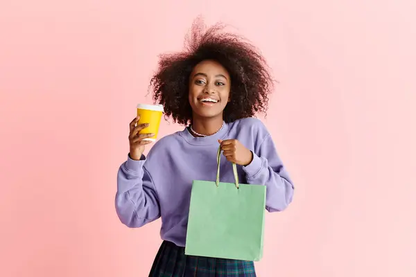 Une femme afro-américaine élégante avec des cheveux bouclés tenant une tasse et un sac en papier dans un cadre de studio. — Photo de stock