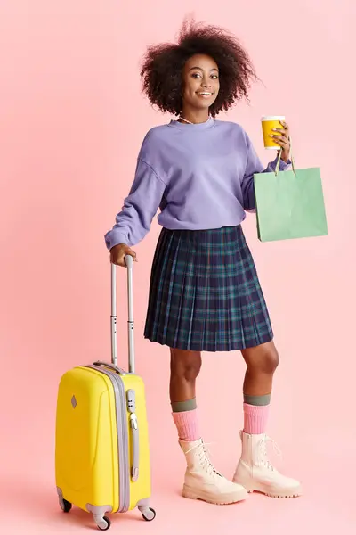 Giovane donna afroamericana in maglione viola e gonna a quadri, pronta per viaggiare con una valigia gialla brillante. — Foto stock