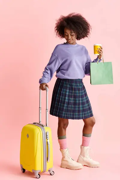 Une belle jeune femme afro-américaine aux cheveux bouclés dans un pull violet et jupe à carreaux, portant une valise jaune. — Photo de stock
