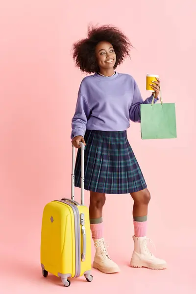 Una giovane donna afroamericana con i capelli ricci tiene una tazza di caffè e una valigia, alla moda e pronta a viaggiare. — Foto stock