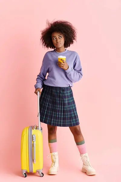 Una giovane donna afroamericana con i capelli ricci in un maglione viola e gonna a quadri, che tiene una valigia gialla e una tazza di caffè in un ambiente da studio. — Foto stock