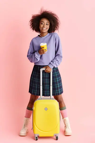 Una hermosa joven afroamericana con el pelo rizado sostiene una maleta amarilla y una taza de café, lista para viajar. - foto de stock