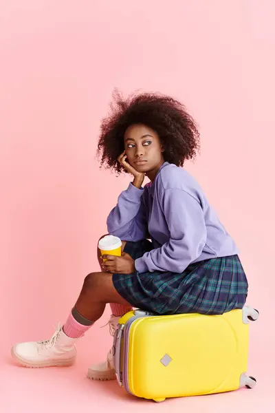 Una joven afroamericana aburrida con el pelo rizado se sienta encima de una maleta amarilla, perdida en el pensamiento. - foto de stock