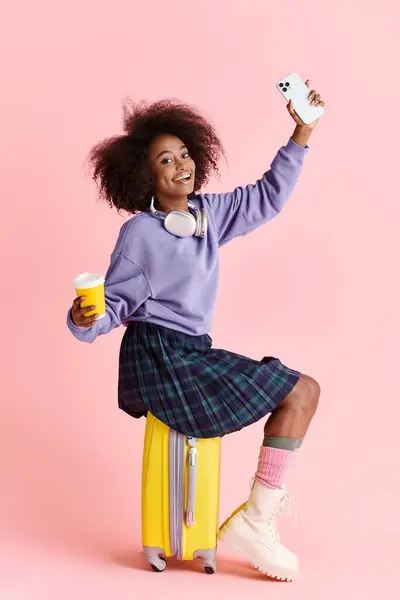 Una hermosa joven afroamericana con el pelo rizado se sienta en una maleta, sosteniendo una taza de café. - foto de stock