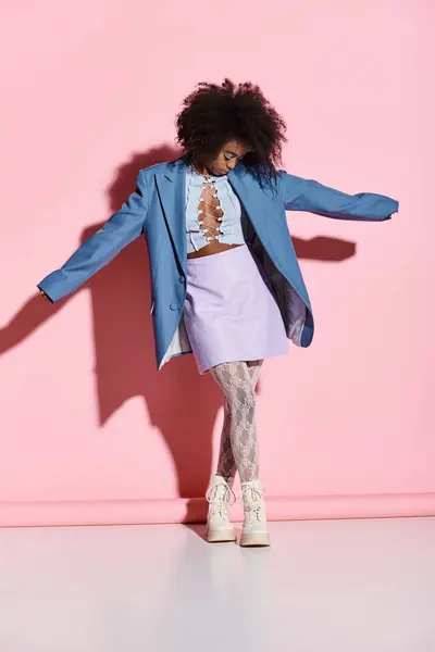 A stylish young African American woman with curly hair stands confidently in front of a vibrant pink wall in a trendy studio setting. — Stock Photo
