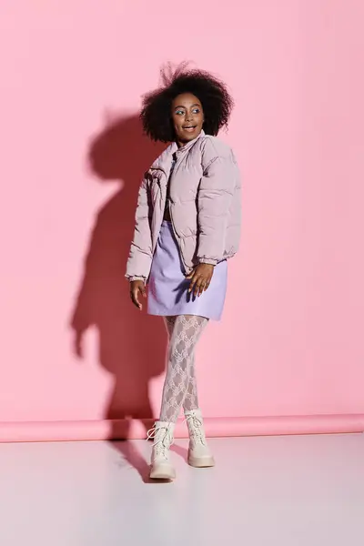 A stylish young African American woman with curly hair stands confidently in front of a vibrant pink wall. — Stock Photo
