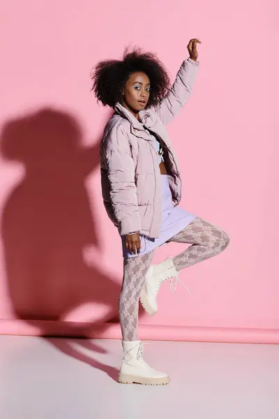 A stylish young African American woman with curly hair standing gracefully in front of a vibrant pink wall. — Stock Photo