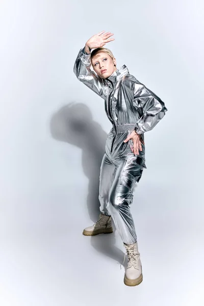 Atractiva mujer en traje futurista plateado posando en movimiento sobre fondo gris y mirando hacia otro lado - foto de stock