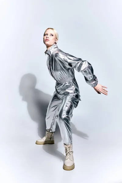 Atractiva mujer en traje futurista plateado posando en movimiento sobre fondo gris y mirando hacia otro lado - foto de stock