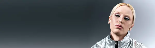Шикарна жінка з світлим волоссям у футуристичному срібному одязі, що позує в русі на сірому тлі, банер — стокове фото