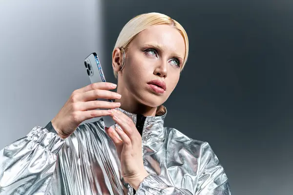 Hermosa mujer en traje de plata futurista posando con el teléfono y mirando hacia otro lado en el fondo gris - foto de stock