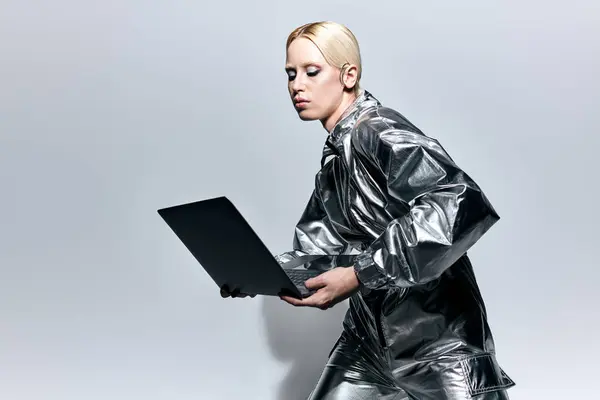 Außergewöhnliche blonde Frau mit Make-up in futuristischen Klamotten schaut auf ihren Laptop vor grauem Hintergrund — Stockfoto