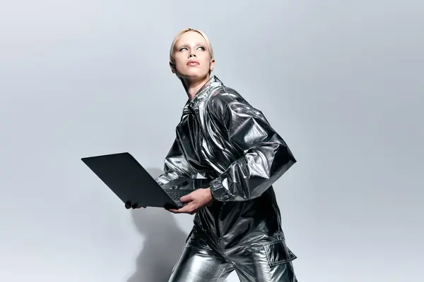 Mujer hermosa extravagante en ropa robótica plateada posando en movimiento con el ordenador portátil y mirando hacia otro lado - foto de stock