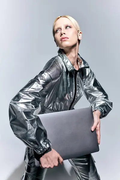 Mujer atractiva extravagante en la ropa robótica de plata posando en movimiento con el ordenador portátil y mirando hacia otro lado - foto de stock