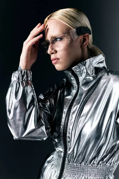 Atractiva mujer rubia con gafas de ciencia ficción en traje futurista mirando hacia otro lado en el fondo gris oscuro - foto de stock
