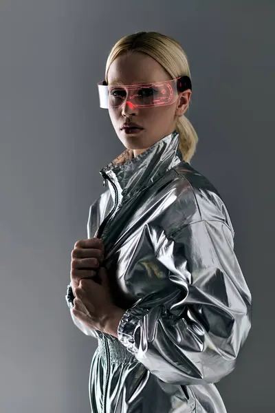 Привлекательная блондинка в футуристических очках в серебряном наряде, смотрящая в камеру на сером фоне — стоковое фото