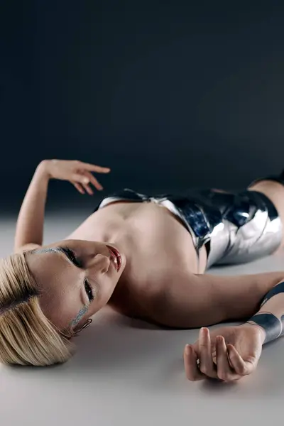 Bizarro hermosa modelo femenina con maquillaje y cabello rubio en ropa futurista tumbado en el suelo - foto de stock