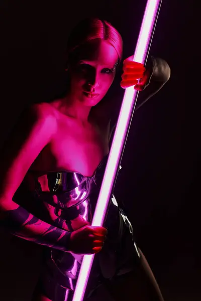 Mujer peculiar en traje futurista metálico sosteniendo palo de lámpara led rosa y mirando a la cámara - foto de stock