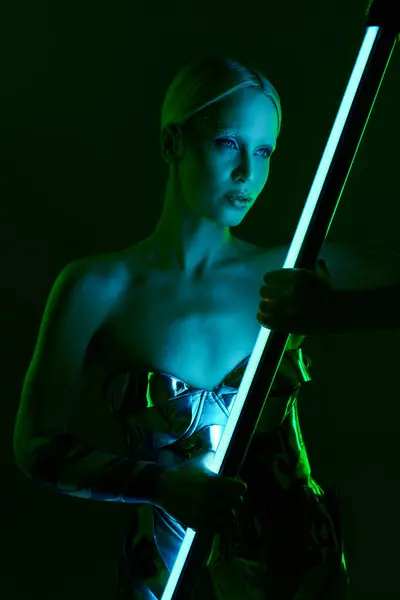 Extraordinaria mujer en traje robótico metálico sosteniendo palo de lámpara led azul y mirando hacia otro lado - foto de stock