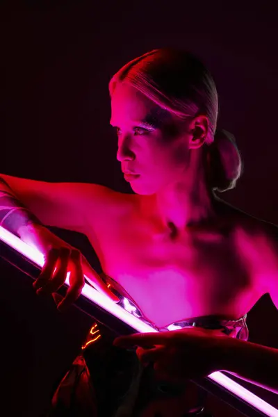 Atractiva mujer peculiar en traje futurista metálico sosteniendo palo de lámpara led rosa y mirando hacia otro lado - foto de stock