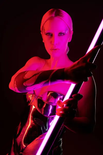 Mujer peculiar en traje futurista metálico sosteniendo palo de lámpara led rosa y mirando a la cámara - foto de stock