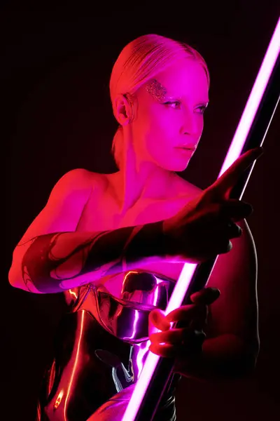 Atractiva mujer en traje futurista metálico sosteniendo palo de lámpara led rosa y mirando hacia otro lado - foto de stock