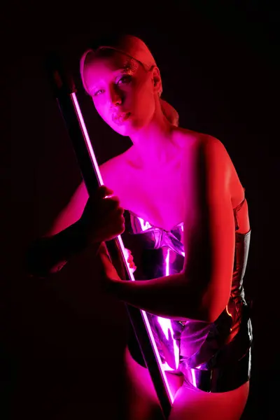Atractiva mujer en traje futurista metálico sosteniendo palo de lámpara led rosa y mirando a la cámara - foto de stock