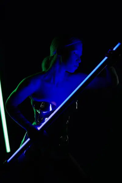 Bella donna particolare in argento abbigliamento robotico in posa con bastone lampada LED blu e guardando altrove — Foto stock