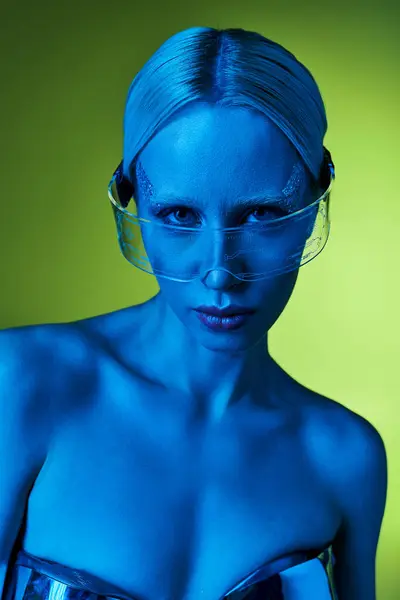 Причудливая женщина с светлыми волосами и фантастическими очками в синем свете на зеленом фоне, смотрящая в камеру — стоковое фото