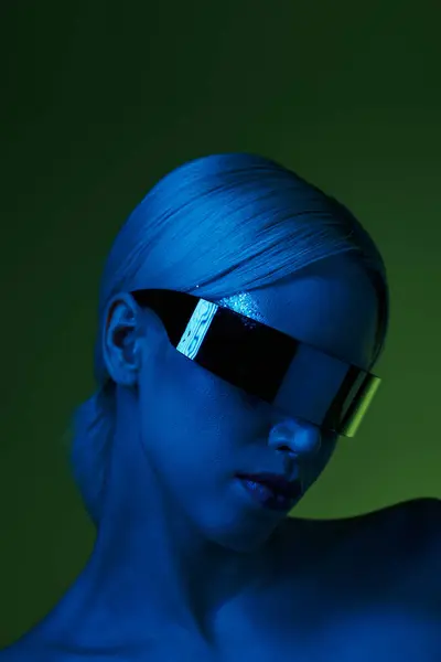 Femme blonde extravagante avec des lunettes de soleil robotiques science-fiction posant dans des lumières bleues sur fond vert — Photo de stock