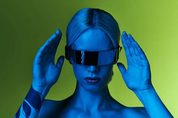 Futuristic blonde woman with sci fi robotic sunglasses posing in blue lights on green backdrop — Stock Photo