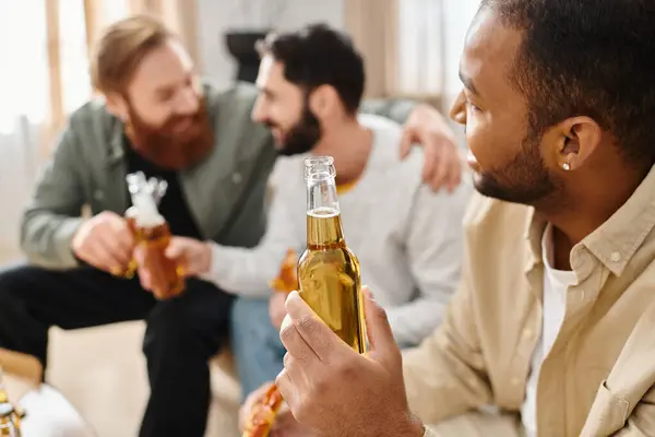 Tres hombres alegres e interraciales en atuendo casual sentados alrededor de una mesa, pegados a las cervezas y disfrutando de un buen rato juntos. - foto de stock