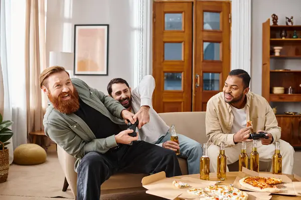 Три веселых человека разных национальностей сидят за столом, смеясь и наслаждаясь пиццей и пивом в непринужденной обстановке дома. — стоковое фото