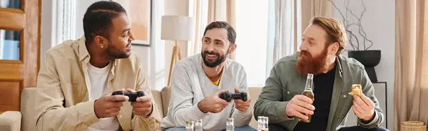 Tre allegri uomini interrazziali si siedono attorno a un tavolo con telecomandi, condividono risate e cameratismo in un ambiente casual. — Foto stock