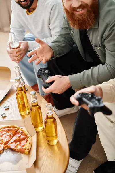 Deux hommes engagés dans des jeux vidéo, tout en dégustant de la bière dans un cadre décontracté à la maison, accompagnés de leur ami interracial. — Photo de stock